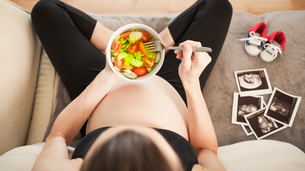 pregnant person eating salad next to sonogram photos and baby shoes